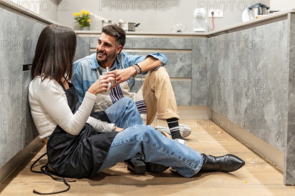 Two chefs are sitting on the floor of a modern kitchen, taking a break and chatting while enjoying a cup of coffee