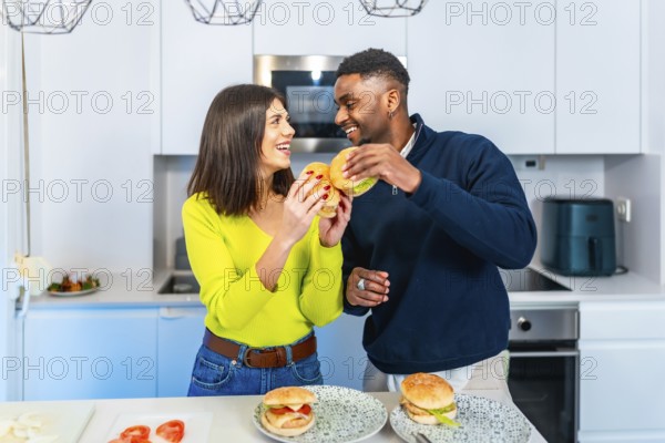 Cheerful couple enjoying a meal together, holding burgers and smiling in their contemporary kitchen
