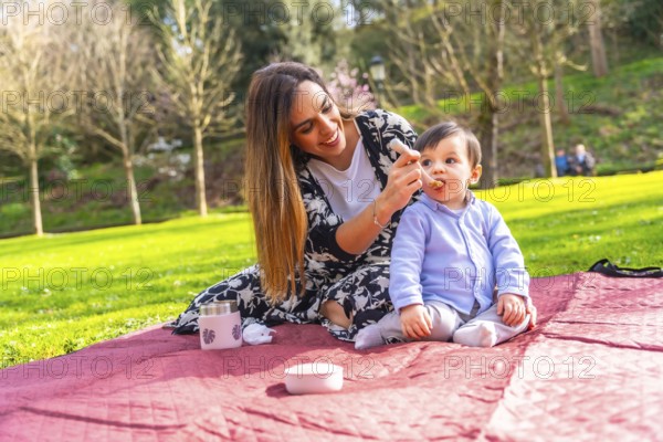 Hispanic mother feeding her baby son solid food with a spoon while having picnic in a park, enjoying family time together