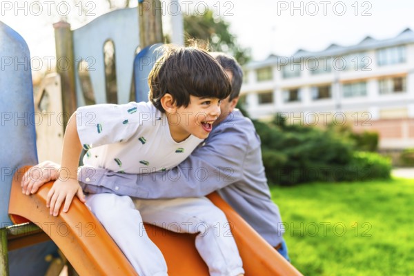 Grandfather holding grandson sliding down an orange slide at playground, having fun together on a sunny day