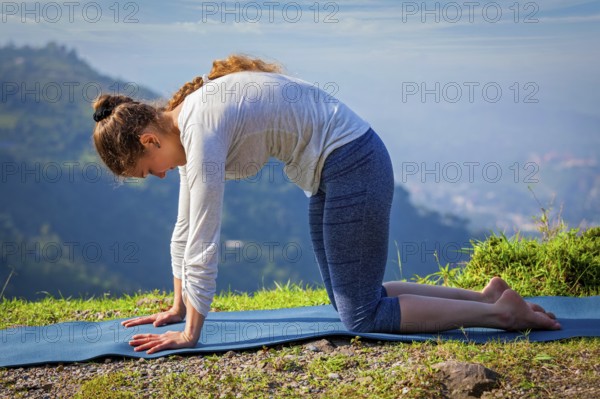 Yoga exercise outdoors, sporty fit woman practices yoga asana Marjariasana, cat pose outdoors in Himalayas