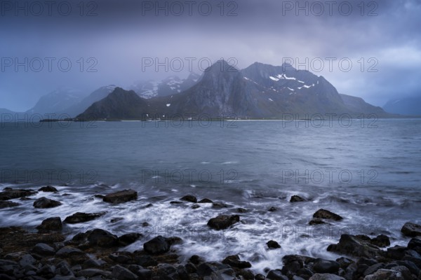 Landscape with sea and mountains on the Lofoten Islands, view across the fjord to the small town of Flakstad and the mountain Flakstadtinden as well as other mountains. Rocks in the foreground. At night at the time of the midnight sun, clouds in the sky, bad weather. Long exposure, early summer. Flakstadoya, Lofoten, Norway, Europe