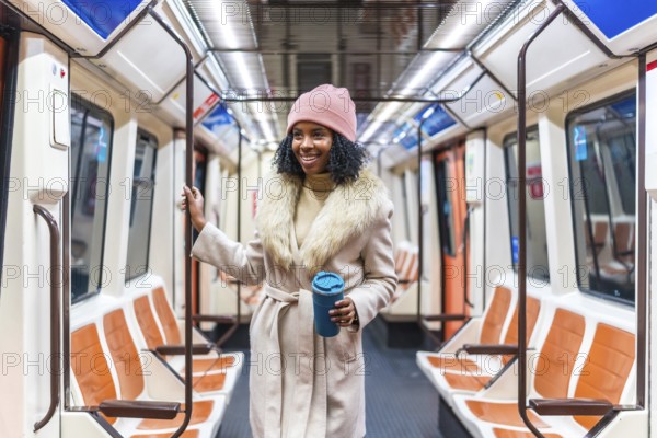 Smiling young black woman wearing winter coat and pink woolen hat holding reusable coffee cup standing on subway train