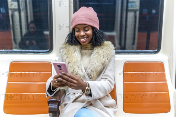 Smiling young black woman wearing winter clothes using smartphone while sitting on subway train