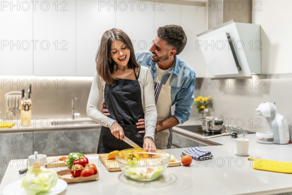 Young smiling couple wearing aprons preparing a healthy vegetable salad together in their modern kitchen