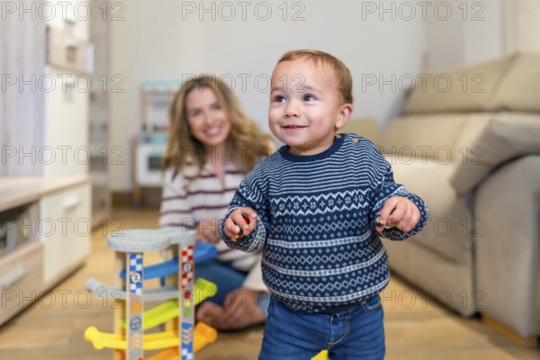 Happy baby boy learning to walk in living room with his mother watching and smiling, playing with a toy car ramp