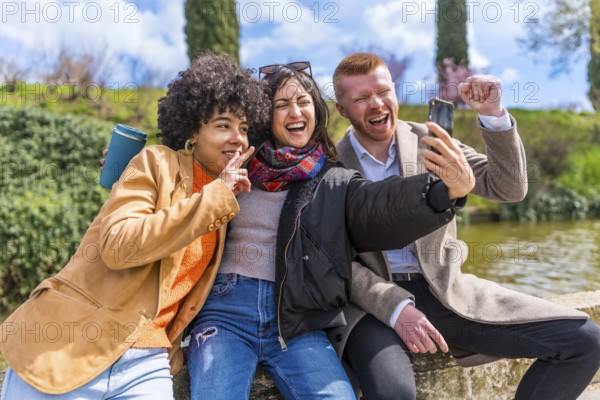 Three cheerful multi ethnic friends are making a selfie while sitting on a wall in a park next to a lake