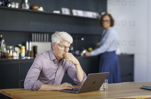 Symbolic photo on the subject of division of labour for couples in the household. A man sits in a kitchen at his laptop while a woman washes dishes in the background. Berlin, 13.08.2024