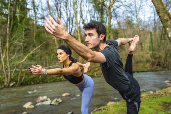 Yoga instructors performing a balancing pose by a river, showcasing flexibility and mindfulness in a natural setting