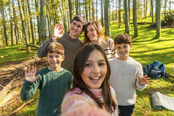 Caucasian family taking selfie and waving at camera standing in an alpine forest enjoying nature together in a sunny day