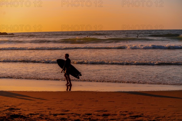 A man is walking on the beach with a surfboard. The sky is orange and the water is calm