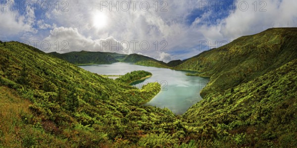 A clear lake surrounded by green hills and mountains under a blue sky with clouds on a sunny day, crater lake Lagoa do Fogo, Caldeiras, Sao Miguel Island, Azores, Portugal, Europe