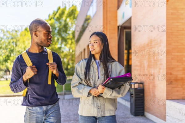 Two african and chinese multi-ethnic friends talking walking along university campus carrying folders and notebooks