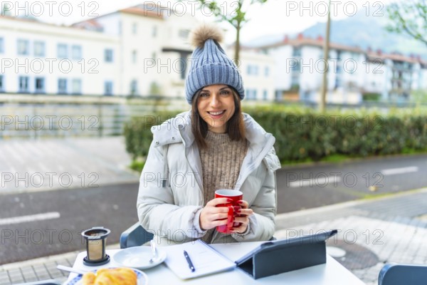 Beauty female caucasian entrepreneur working with digital tablet sitting in an outdoor cafeteria and warming hands with hot tea in a cold winter day
