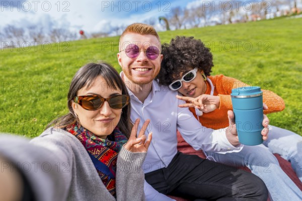 Three happy multi ethnic friends are taking a selfie in a park, gesturing victory signs and showing a reusable coffee cup