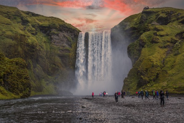 Group of tourists looking at the beautiful rainbow at the impressive Skogarfoss waterfall in southern Iceland, photo taken in summer on a beautiful sunset