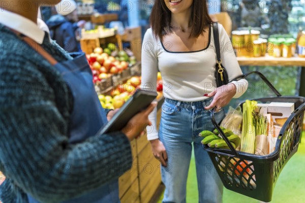 Saleswoman using digital tablet assisting customer carrying shopping basket with fresh produce in organic supermarket