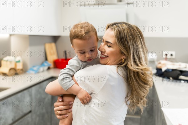 Smiling mother holding her baby son in a home kitchen, enjoying a tender moment of affection and bonding