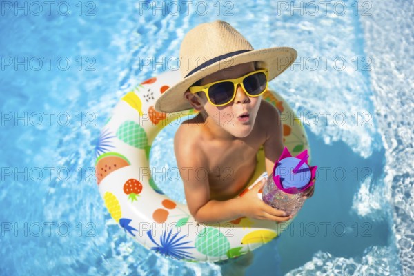 Top view of an expressive caucasian boy wearing sunglasses and sun hat playing inside a pool with rubber ring float