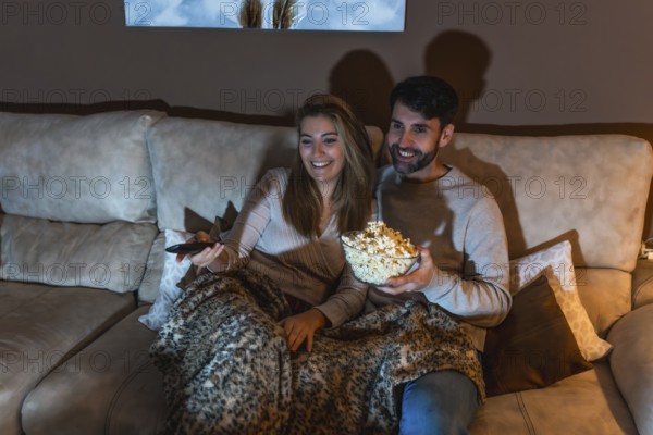Young couple enjoying a movie night at home, sharing popcorn and relaxing on the sofa under a blanket