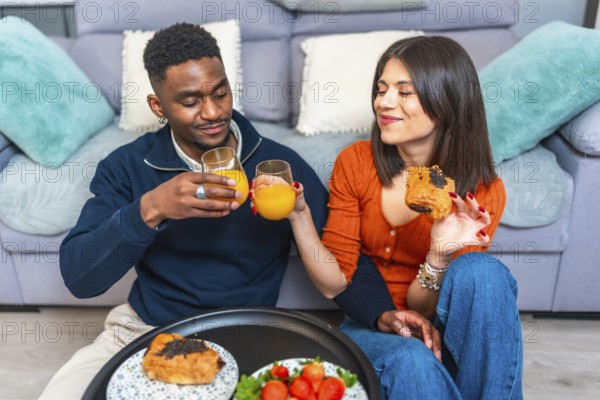 Multiethnic couple sits together, enjoying breakfast and wine, creating a cozy and intimate dining experience in their living room