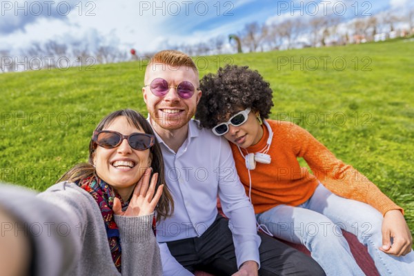 Three multi ethnic friends are sitting on the lawn in a park, taking a selfie and waving at the camera