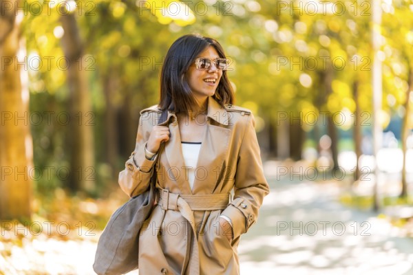 A woman wearing a tan trench coat and sunglasses walks down a sidewalk. She is smiling and she is enjoying her day. Autumn or winter concept in the city