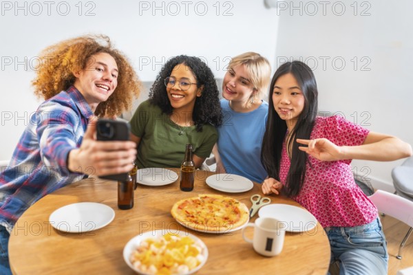 Four diverse friends are taking a selfie with a smartphone while enjoying lunch with pizza and beer at home