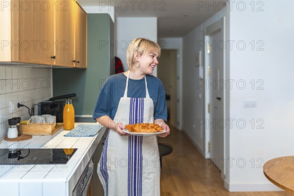 Smiling woman wearing apron carrying plate of fresh croissants in contemporary kitchen, enjoying homemade breakfast