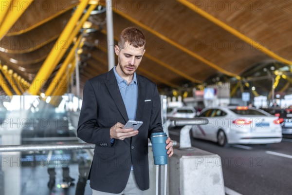 Young businessman using smartphone and holding reusable coffee cup while waiting for taxi or rideshare at airport