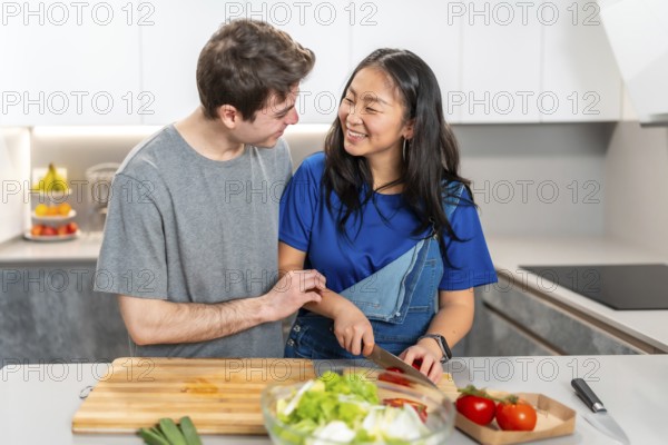 Multiethnic couple sharing a joyful moment while preparing a meal in a sleek, modern kitchen. Fresh vegetables and a cutting board are in view