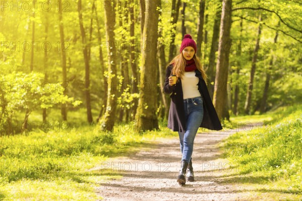 Latin girl with a black jacket, scarf and red woolen hat enjoying in an autumn park, walking along a path with a coffee at sunset