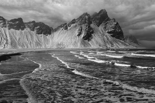 White foamy waves on the beach of Stokkness, behind snowy rocky slopes of Vestrahorn, black and white photo, near Höfn, Sudausturland, Iceland, Europe