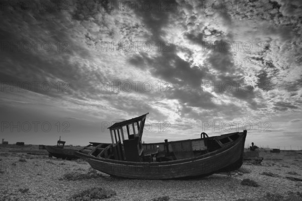 Broken discarded fishing boats on a shingle beach against a dramatic cloudy backdrop, boat graveyard, black and white, Dungeness, Kent, England, United Kingdom, Europe