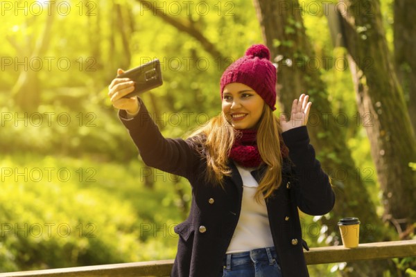Young man in a black jacket, scarf and red woolen hat enjoying in an autumn park, waving on a video call with the phone