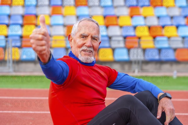 Proud senior man gesturing with thumbs up sitting on a running track after running