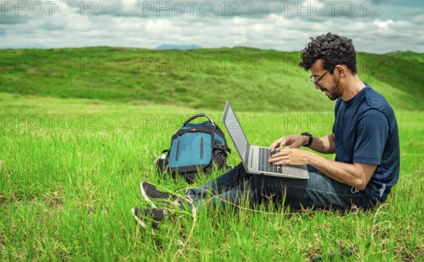 Close up of man sitting on green grass working with his laptop. Smiling freelancer guy working from the field. Smiling man with laptop sitting on the grass in the field