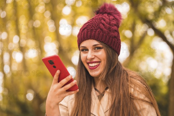 Portrait of a beautiful woman in autumn next to a forest in nature smiling sending a voice note with the phone