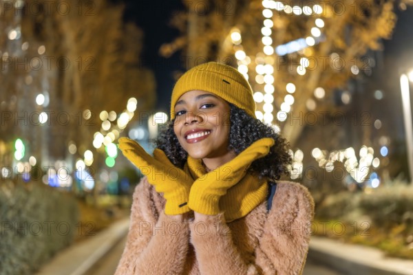 Portrait of a cheerful young woman wearing yellow woolen gloves and hat enjoying christmas lights in the city at night