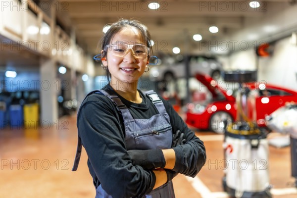 Confident young female mechanic wearing safety glasses and earplugs, smiling with crossed arms in a busy car repair shop