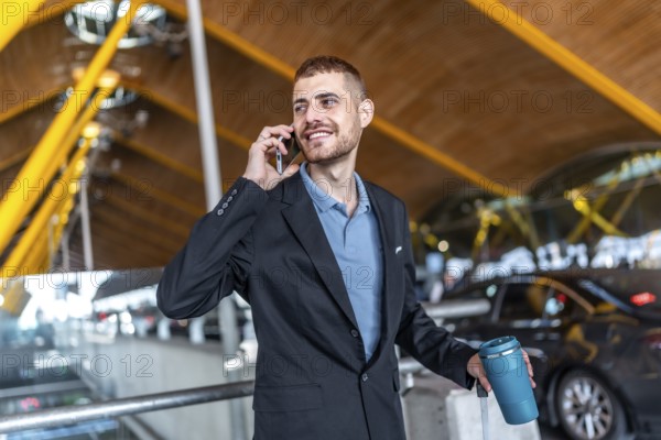 Young businessman smiling while talking on phone, holding his luggage and coffee thermos, at the airport