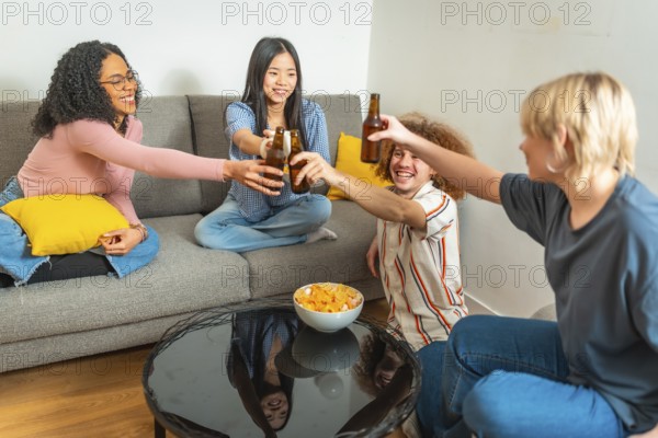 Group of young multi ethnic people having fun and toasting with beer bottles while sitting on a sofa and eating chips at home