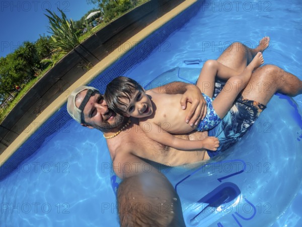 Father and son are taking a selfie while relaxing on an inflatable ring in a swimming pool at a water park during a sunny day