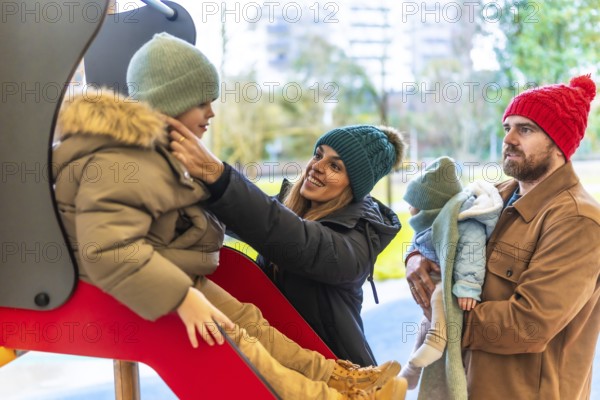 Smiling mother helping her son going down the slide, while father holding baby, enjoying a winter day at the playground