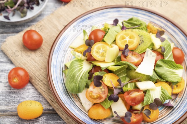Vegetarian salad of pac choi cabbage, kiwi, tomatoes, kumquat, microgreen sprouts on gray wooden background and linen textile. Top view, close up, selective focus
