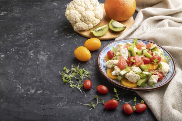 Vegetarian salad of cauliflower cabbage, kiwi, tomatoes, microgreen sprouts on black concrete background and linen textile. Side view, copy space
