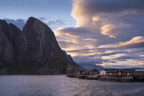 Cloudy landscape, view of Toppoya (Toppoy), the sea and Festhaeltinden mountain. Typical red wooden houses on wooden piles (Rorbuer) . Several coloured lenticular clouds (lenticularis) in the blue sky. At night at the time of the midnight sun in good weather. Early summer. Moskenesoya, Lofoten, Norway, Europe