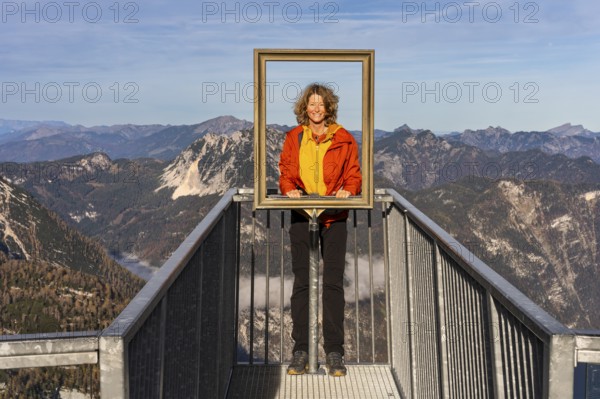 A hiker stands on the 5fingers viewpoint and looks through the picture frame. View from the Dachstein Krippenstein. Autumn, good weather. Some clouds in the mountains. Lake Hallstatt, Salzkammergut, Upper Austria, Austria, Europe