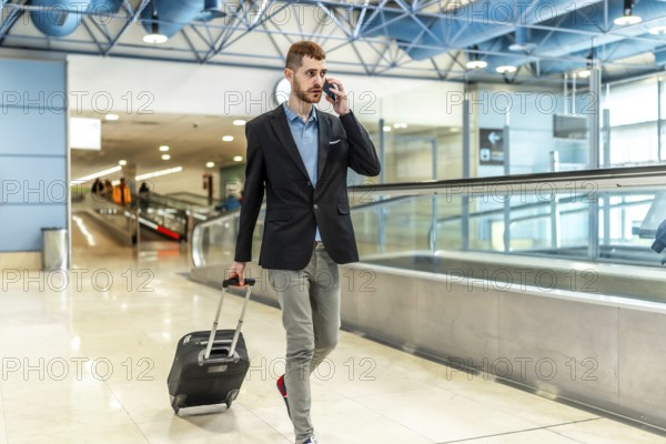 Businessman pulling a suitcase while talking on the phone, confidently walking through a bustling airport terminal