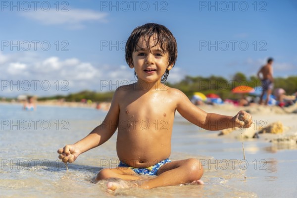 Happy child playing with sand sitting on the shore of a paradisiacal beach with crystalline waters and other tourists in the background